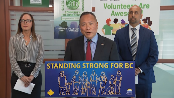 Deux hommes et une femme donnant une conférence de presse derrière un panneau "standing strong for BC".