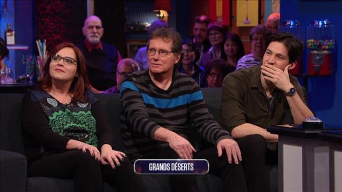 Une femme et deux hommes sur le plateau d'un jeu télévisé, devant une petite foule en arrière-plan.