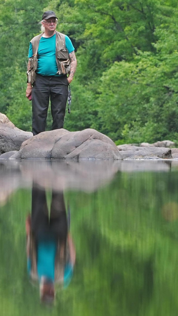 Un homme en habit de pêche se tient debout derrière des rochers dans une rivière, calme, entouré de forêt.