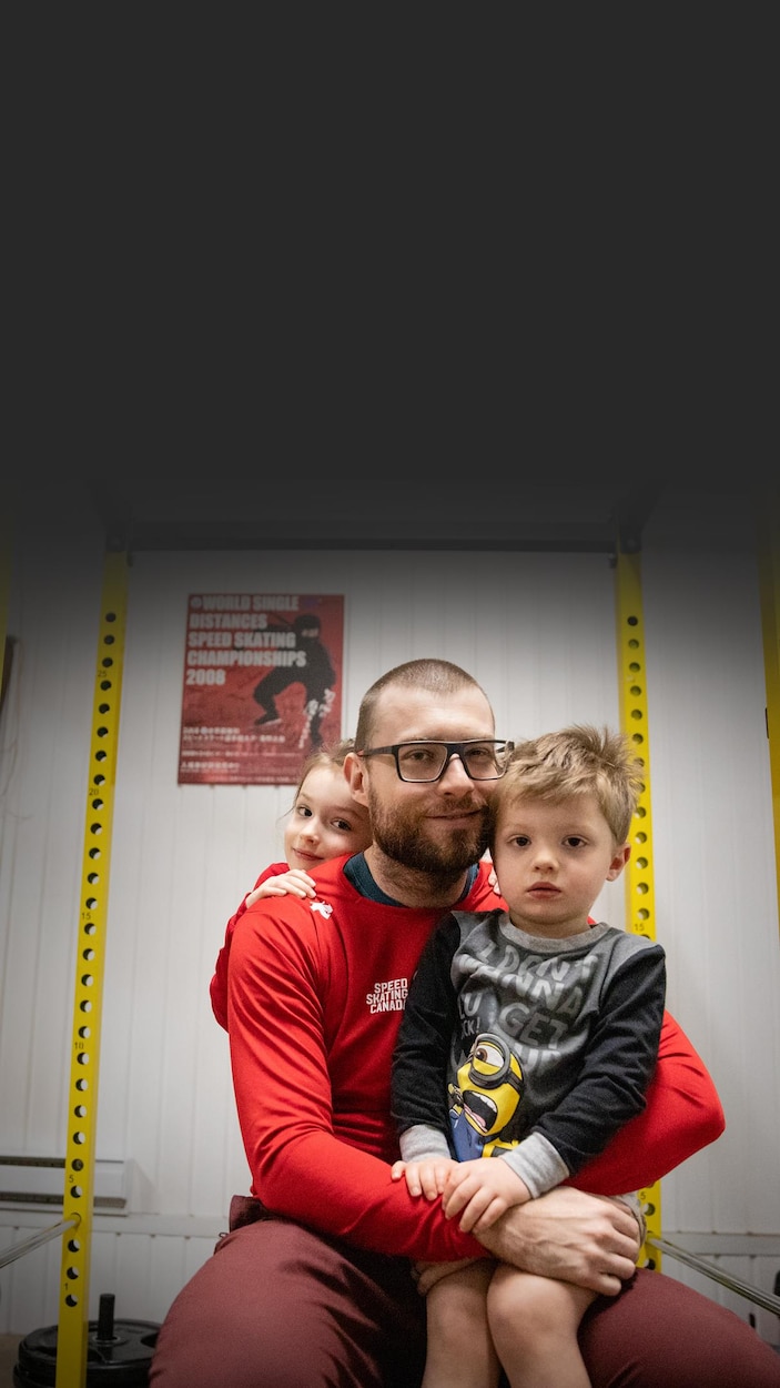 Un homme portant des lunettes et un haut Speed Skating Canada sourit, entouré de deux jeunes enfants dans une salle de sport.