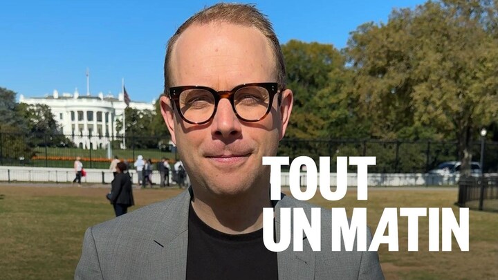 Le journaliste Louis Blouin est debout, il tient un petit micro devant la Maison-Blanche.