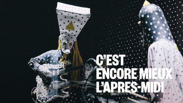 Deux musiciens masqués et vêtus de tenues à pois noirs et blancs performent sur scène. L'un joue d'une guitare double manche sous une coiffe trapézoïdale imposante, tandis que l'autre, de profil, s'installe derrière une batterie devant un fond pointillé.