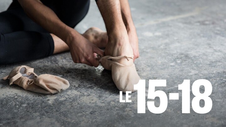 Un danseur est assis et met un chausson de danse sur son pied gauche.