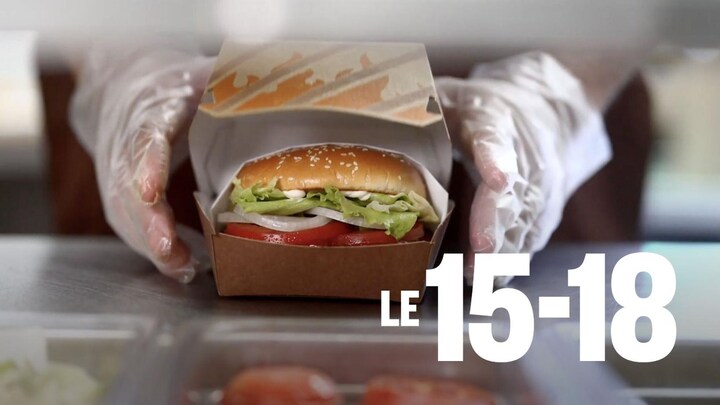 Des mains gantées tiennent un hamburger avec des tomates, des oignons et de la laitue dans une boite en carton.
