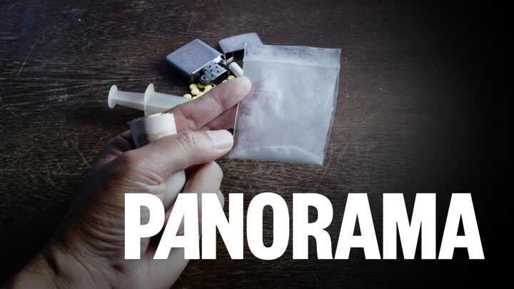 Une main qui tient de l'argent et de la poudre d'héroïne dans un sac. Une seringue, un médicament, une cigarette et un briquet sont déposés sur une table en arrière-plan.