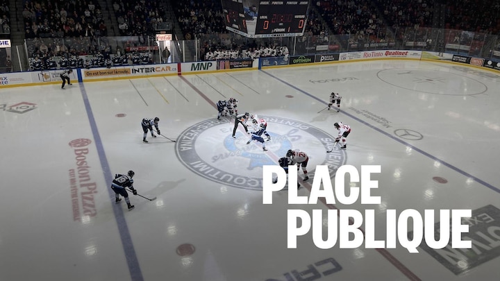 Le début d'un match de hockey au Centre Georges-Vézina de Saguenay. Deux équipes, vêtues de bleu et de rouge/blanc, se font face sur la patinoire pour une mise au jeu devant une foule dense.