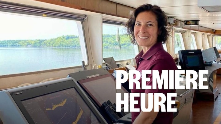 Une femme capitaine se tient au centre du pont d'un navire, vêtue d'un polo marron à manches courtes et d'une jupe foncée. Elle regarde vers la caméra avec un sourire confiant, ses mains posées sur les commandes devant elle.