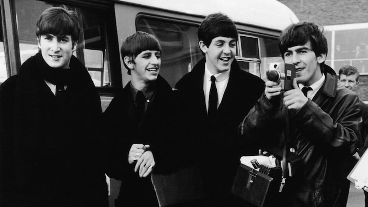 Les Beatles sont près d'un autobus et sourient, alors que George Harrison tient une caméra.