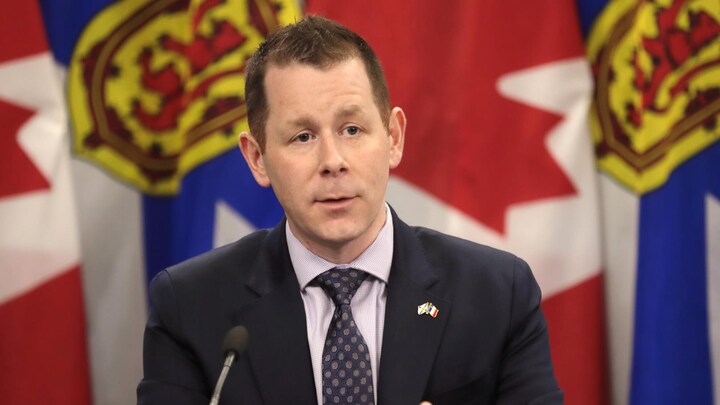 Un politicien en complet parle, assis à une table devant des drapeaux de la Nouvelle-Écosse et du Canada.