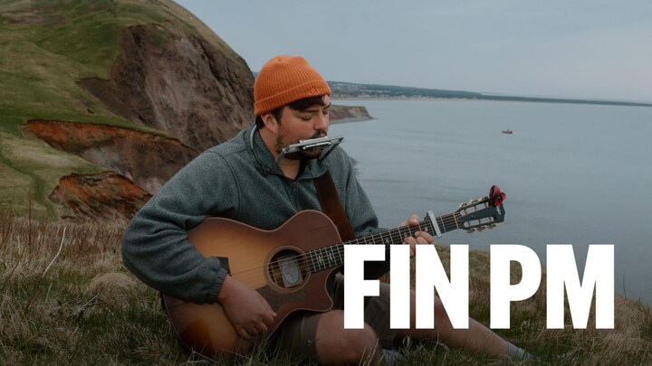 Émile Bilodeau assis dans l'herbe sur une bute au bord de l'eau avec sa guitare et son harmonica.