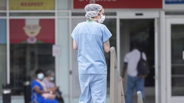 Une infirmière en uniforme, portant un bonnet et un couvre-visage, se tient debout devant les urgences d'un hôpital.