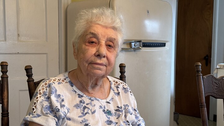 Jeannette Mercier, une femme âgée, regarde l'objectif, assise dans sa cuisine, devant un frigo d'époque.