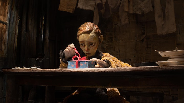 Une marionnette représentant une fille déballe un cadeau dans une cuisine vétuste.