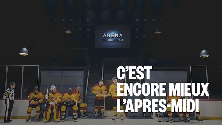 Scène de spectacle se déroulant dans un aréna de hockey. Des comédiens vêtus d'uniformes de hockey jaunes et rouges sont assis sur le banc des joueurs. Deux joueurs sont debout au centre, et un arbitre est à gauche. Un panneau lumineux au-dessus indique "ARÉNA R. GOUDREAU".