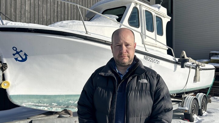 Un homme regarde la caméra, un bateau de pêche au homard derrière lui.