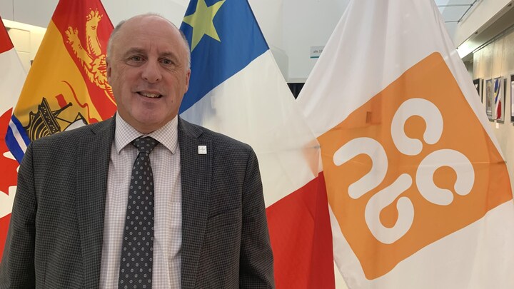 Un homme sourit à la caméra, devant les drapeaux du Nouveau-Brunswick, de l'Acadie et du CCNB