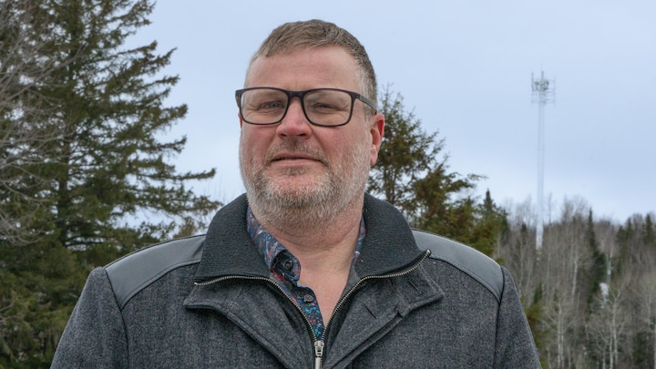 Le maire regarde la caméra. Derrière lui, il y a une tour cellulaire qui est sur une montagne derrière lui.