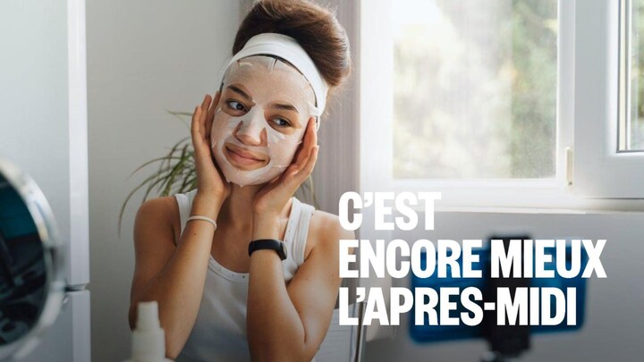A girl poses in front of her phone with a sheet mask on her face.