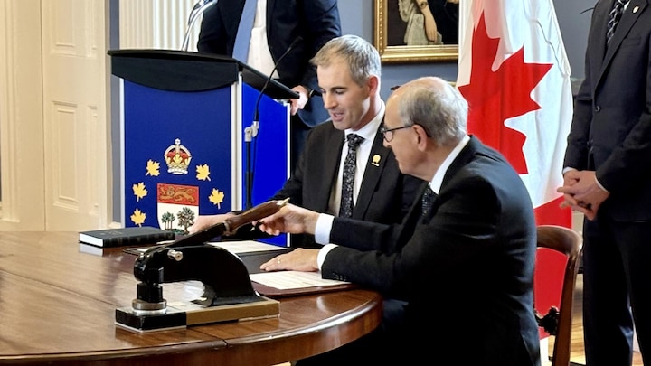 Le nouveau ministre est assis à une petite table ronde à côté du lieutenant-gouverneur, qui lui tend un stylo.