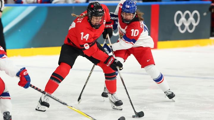 Une joueuse de hockey sur glace protège la rondelle avec son bâton lors d'un match olympique. Elle est poursuivie de près par une joueuse de l'équipe de adverse.