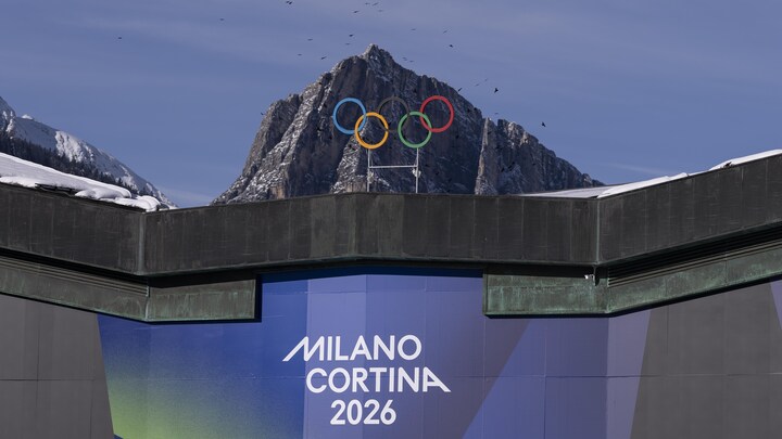 Le logo des Jeux de Milan-Cortina avec les anneaux olympiques est placé devant une montagne enneigée.