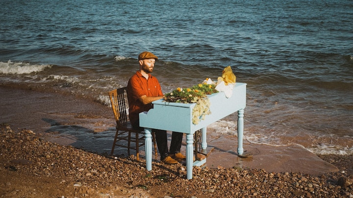 Un homme portant chemise orangée et casquette joue du piano sur la grève. Des fleurs sont placées sur l'instrument de couleur bleu poudre.