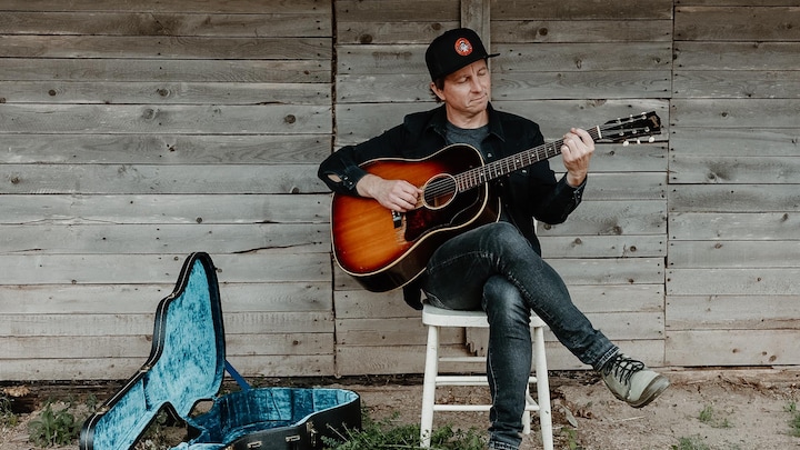 Un homme coiffé d'une casquette joue de la guitare sur une chaise. L'étui de l'instrument, est ouvert; on voit son intérieur en velours bleu.