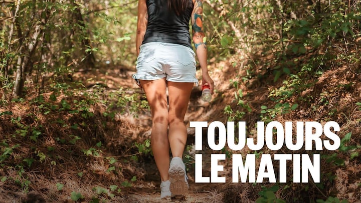 Une femme de dos marchant dans la forêt. Elle tient une bouteille d'eau dans sa main droite.