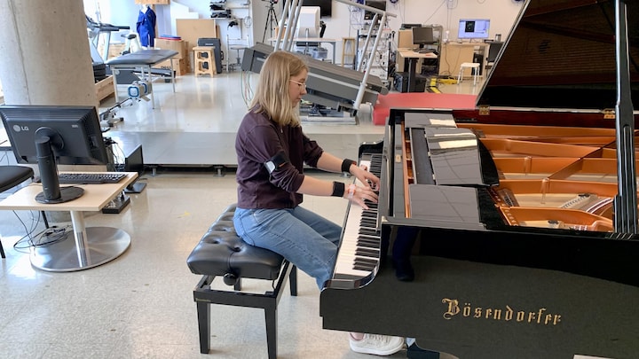 Une pianiste joue une pièce musicale sur un piano à queue, dans une grande salle aux allures de laboratoire. Elle porte de petits instruments aux bras, aux poignets et aux mains.