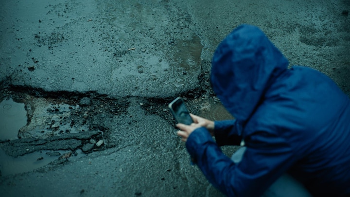 Une personne prend en photograph  un nid-de-poule avec lad   téléphone cellulaire alors qu'il pleut. 