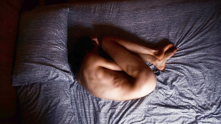 Une personne couchée en position fœtale, nue, dans un lit et vue du haut.
