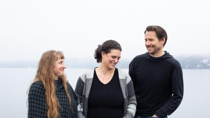 Trois personnes prennent la ose devant un plan d'eau brumeux.