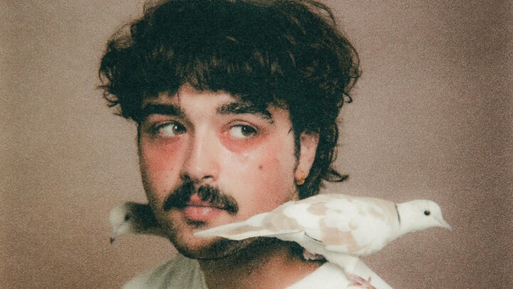 Un homme portant la moustache. Il a les cheveux bruns et des colombes sont posées sur sa tête et ses épaules.