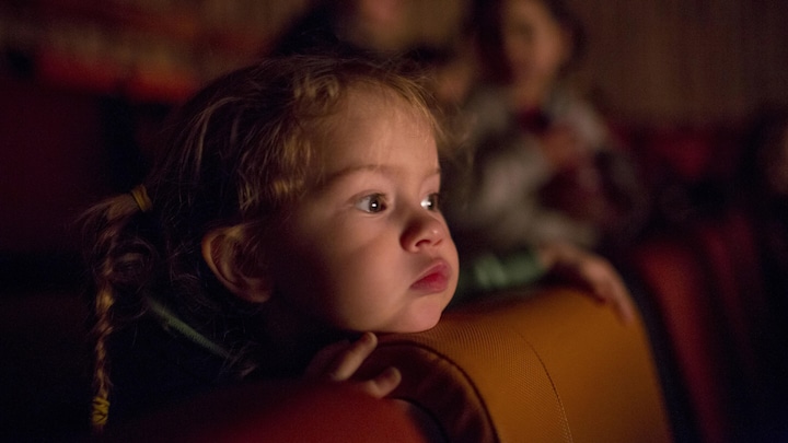 Gros plan du visage d'une petite fille qui regarde un spectacle dans une salle. Sa tête est appuyée sur un siège.