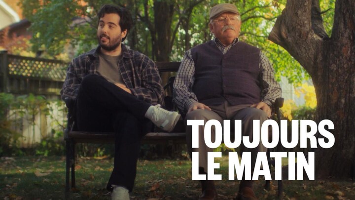 Deux hommes, un jeune et un plus vieux sont assis sur un banc dans une cour, sous un arbre.