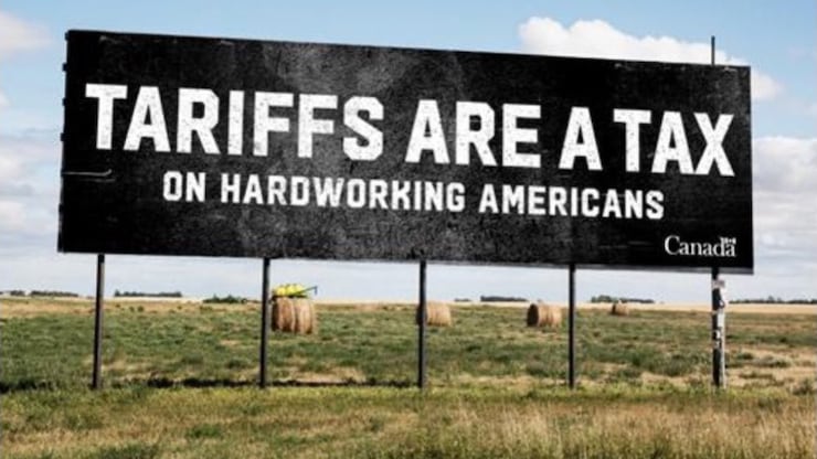 لوحة إعلانية ضخمة سوداء اللون تابعة للحكومة الكندية تم نصبها في حقل زراعي، كُتب عليها: ’’الرسوم الجمركية هي ضريبة على الأميركيين الذين يعملون بجِد‘‘.