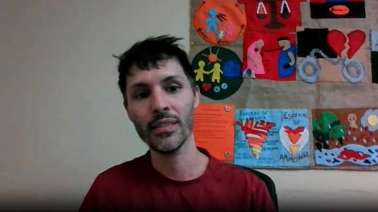 ES: Hombre con barba y camisa roja en videollamada, con un colorido tapiz artesanal en el fondo.
FR: Homme barbu en chemise rouge en appel vidéo, avec une tapisserie artisanale colorée au fond.
EN: Bearded man in a red shirt on a video call, with a colorful handmade tapestry in the background.