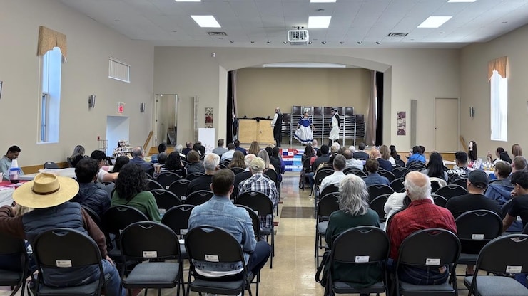 ES: Público sentado en sillas plegables dentro de un salón asistiendo a un evento.
FR: Public assis sur des chaises pliantes dans une salle, assistant à un événement.
EN: Audience seated in folding chairs inside a hall, attending a community event.