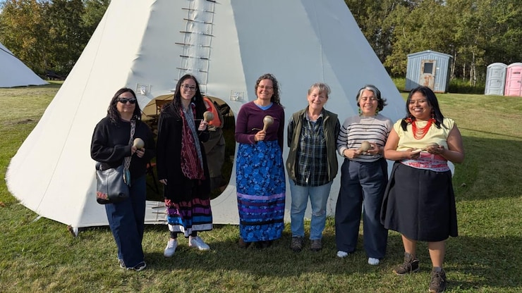ES: Seis mujeres sonrientes sostienen sonajas mientras posan de pie frente a un gran tipi blanco.
FR: Six femmes souriantes tiennent des hochets en posant debout devant un grand tipi blanc en plein air.
EN: Six smiling women holding rattles pose standing together in front of a large white teepee outdoors.