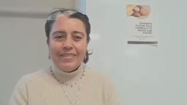 ES: Mujer sonriente con un mechón gris, suéter beige y collar, de pie frente a una pared blanca.
FR: Femme souriante avec une mèche grise et un pull beige, debout devant un mur blanc.
EN: Smiling woman with a gray streak in her hair and a beige sweater, standing by a white wall.