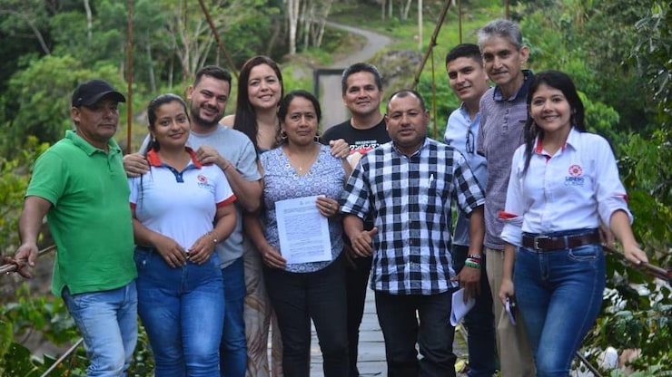 ES: Grupo de nueve personas posando sonrientes sobre un puente colgante de madera en medio de la selva.
FR: Groupe de neuf personnes souriantes posant sur un pont suspendu en bois au milieu de la jungle.
EN: Group of nine smiling people posing together on a wooden suspension bridge surrounded by jungle.