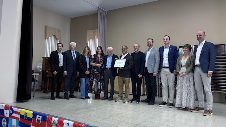 ES: Grupo diverso de personas posa sonriente en un escenario con un diploma enmarcado.
FR: Un groupe diversifié pose en souriant sur une scène avec un diplôme encadré.
EN: A diverse group of people poses smiling on a stage with a framed certificate.