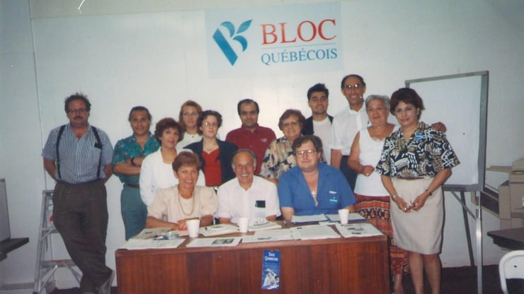 ES: Grupo de personas posando sonrientes tras un escritorio bajo el cartel del "Bloc Québécois".
FR: Groupe de personnes souriantes posant derrière un bureau sous l'affiche du "Bloc Québécois".
EN: Group of smiling people posing behind a desk under a "Bloc Québécois" political party banner.