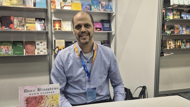 رجل جالس إلى طاولة، وخلفه رفوف مليئة بالكتب.
