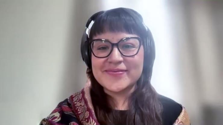 ES: Mujer sonriente con gafas oscuras, flequillo y auriculares grandes sobre una bufanda estampada. FR: Femme souriante avec lunettes, frange et casque audio sur une écharpe colorée. EN: Smiling woman with glasses, bangs, and large headphones over a patterned scarf.