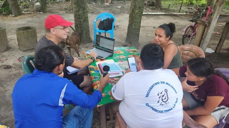 ES: Cinco personas sentadas alrededor de una mesa con mantel verde, una laptop y un micrófono. FR: Cinq personnes sont assises autour d'une table avec un ordinateur portable, un micro et des papiers. EN: Five people are gathered around a table outdoors, looking at a laptop and using a microphone.