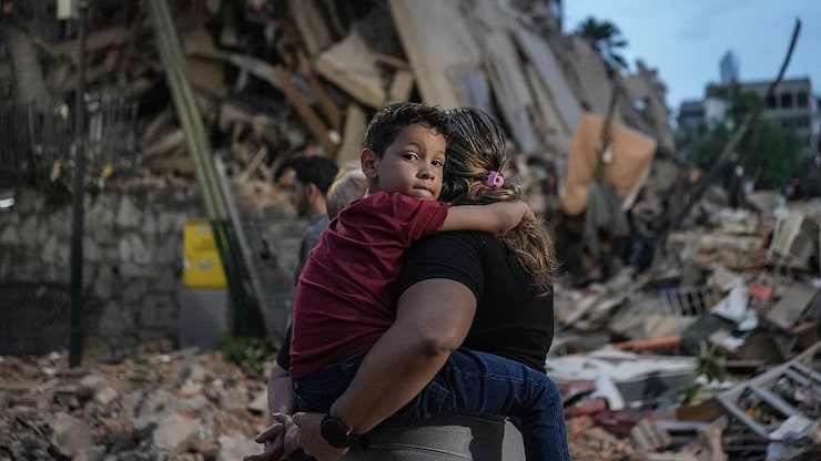 Un enfants dans les bras de sa mère. Dans le fond un immeuble presque totalement détruit.