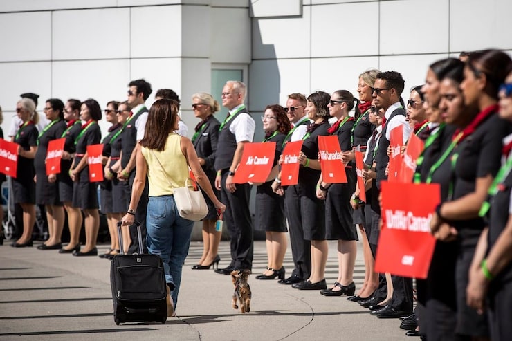 A woman with her luggage and her small dog walk past flight attendants and air personnel standing in line holding signs during a protest in an airport.