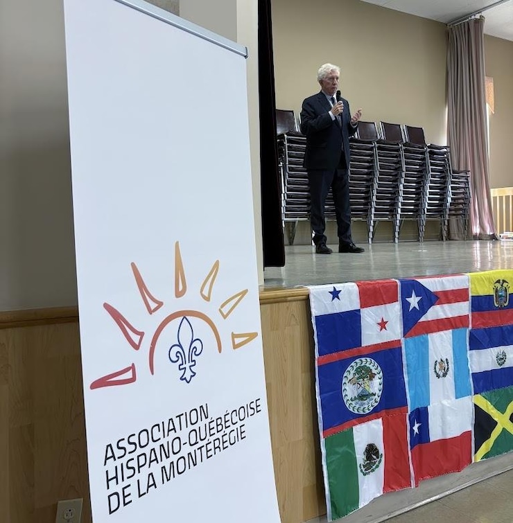 ES: Hombre en traje habla por micrófono en escenario con banderas. Delante, cartel de asociación hispana.
FR: Un homme en costume parle au micro sur une scène avec des drapeaux, derrière une affiche associative.
EN: A man in a suit speaks into a microphone on a stage with flags, behind a Hispanic association banner.