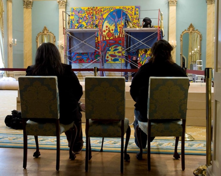 Two women sitting on chairs look at a large painting being installed in an opulent room.
